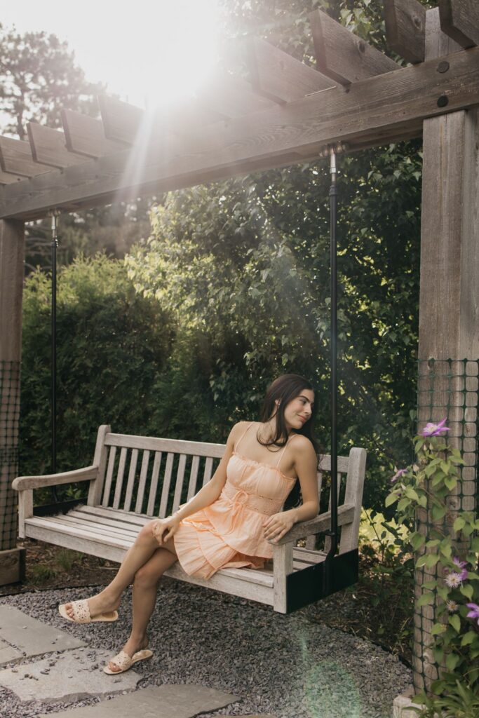 Senior in peach dress sitting on a wooden bench swing under a sunlit pergola