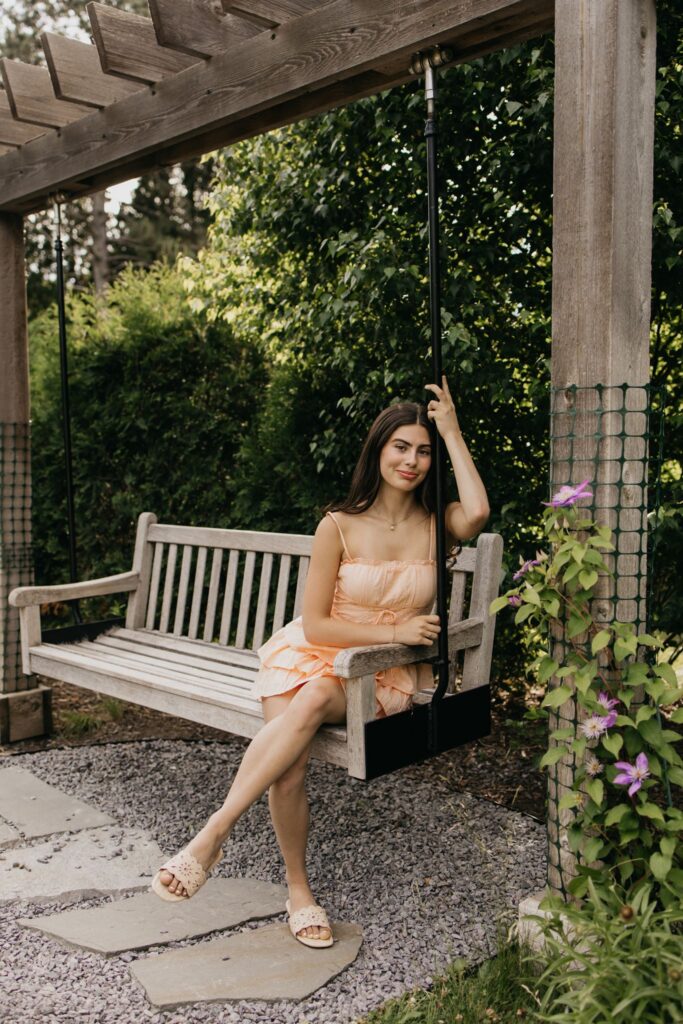 Senior in peach dress sitting on a wooden bench swing under a pergola, full body