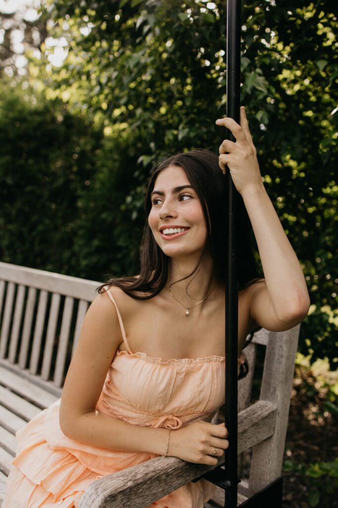 Senior in peach dress sitting on a wooden bench, hand raised on a post, smiling