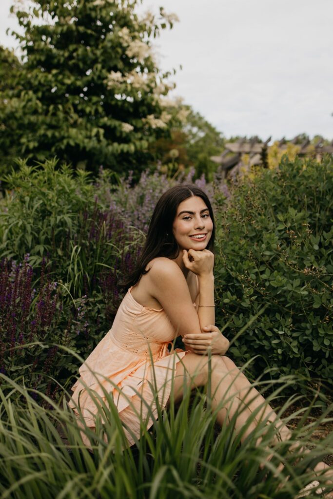 Senior in peach dress sitting in a flower field, chin resting on hand