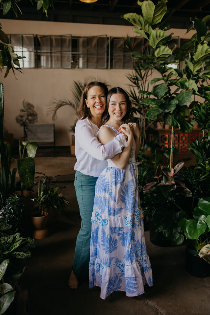Mom in white top laughing with her senior daughter among tall potted plants during Minneapolis senior photography session with mom