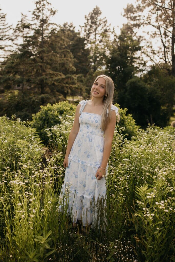 Blonde senior in blue floral maxi dress standing backlit in tall wildflowers at golden hour