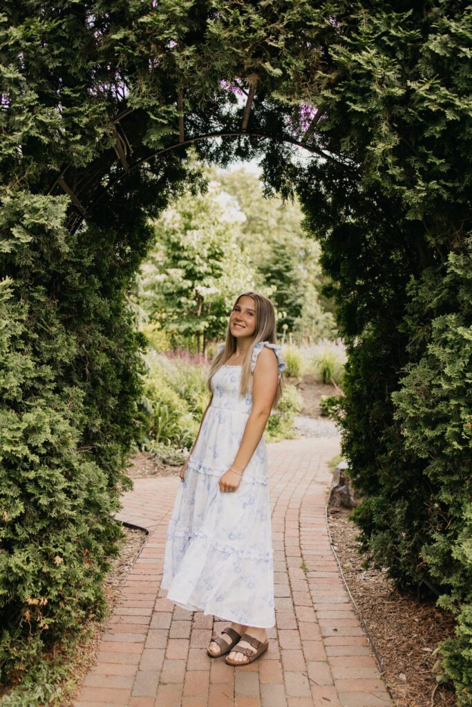 Blonde senior in blue floral maxi dress walking a brick path through a garden archway