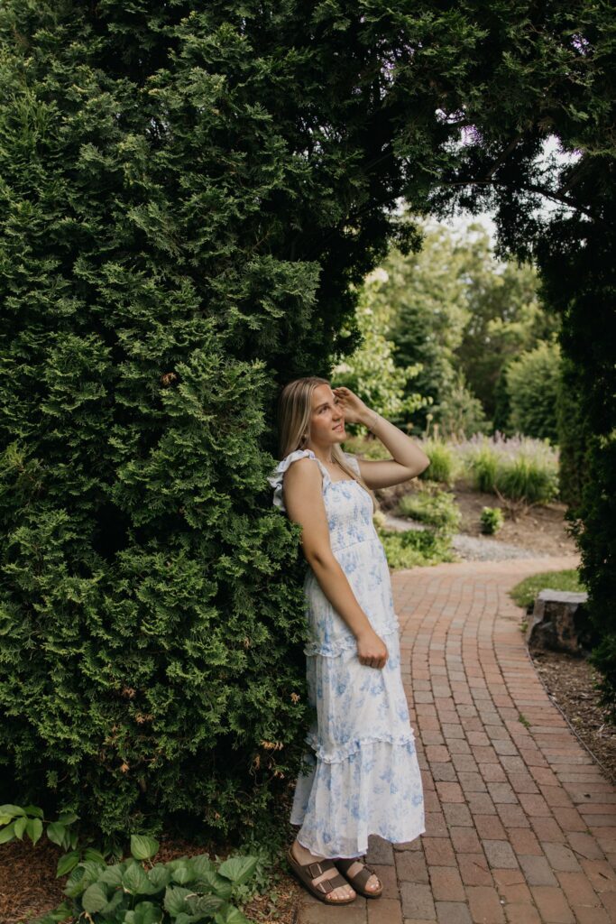 Blonde senior in blue floral maxi dress standing by a hedge on a brick garden path