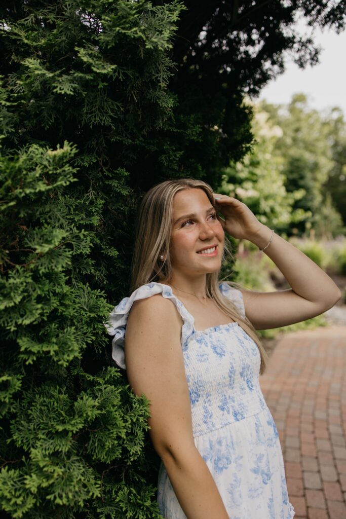 Blonde senior in blue floral dress leaning against a hedge on a brick garden path