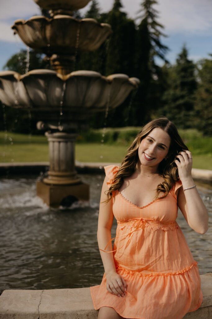 Senior in orange dress sitting on the edge of a tiered garden fountain, hand in her hair