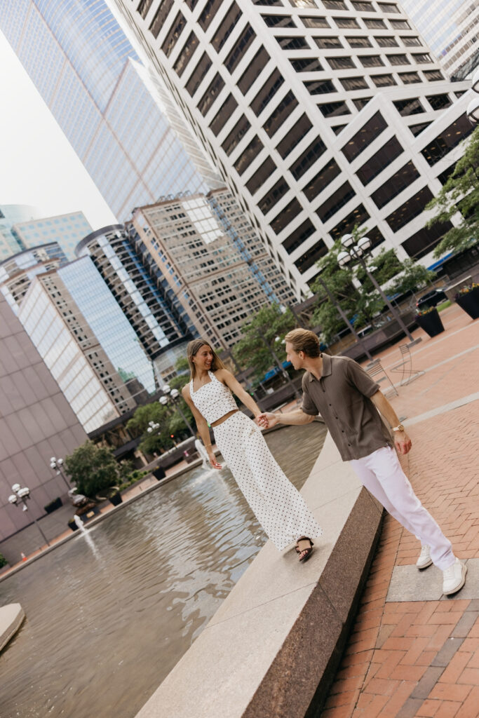 Couple walking along a downtown fountain ledge, city buildings behind them