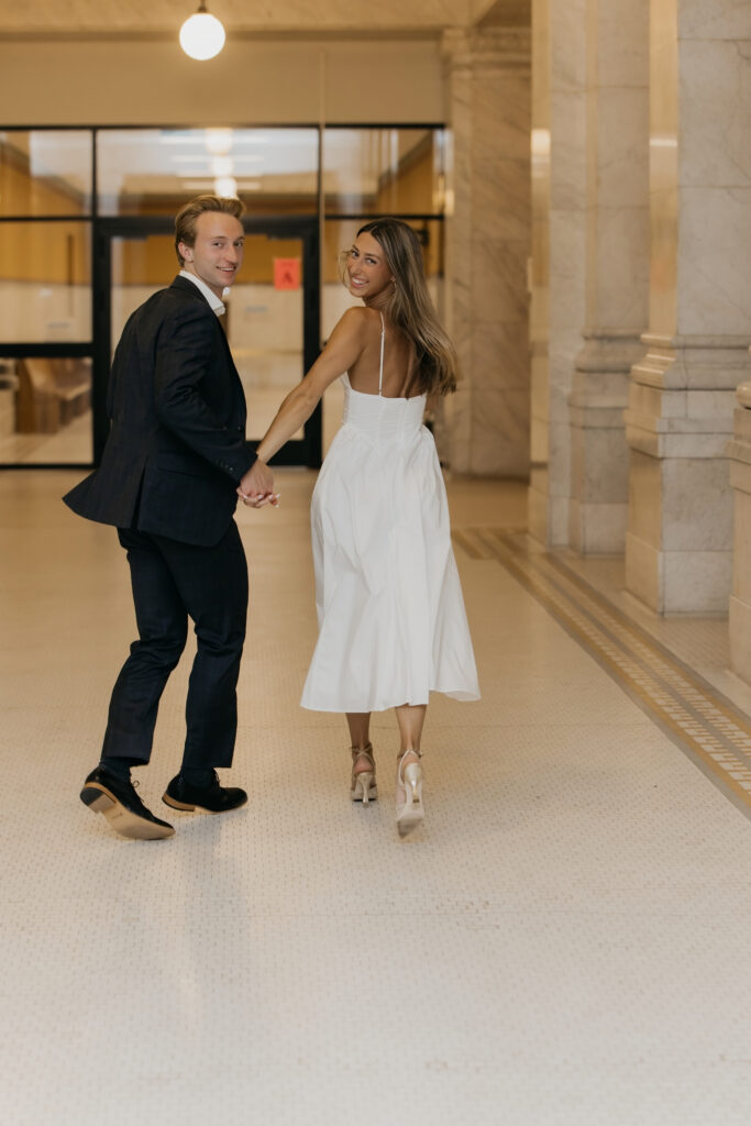 Couple walking hand in hand through a marble building lobby, looking back at the camera