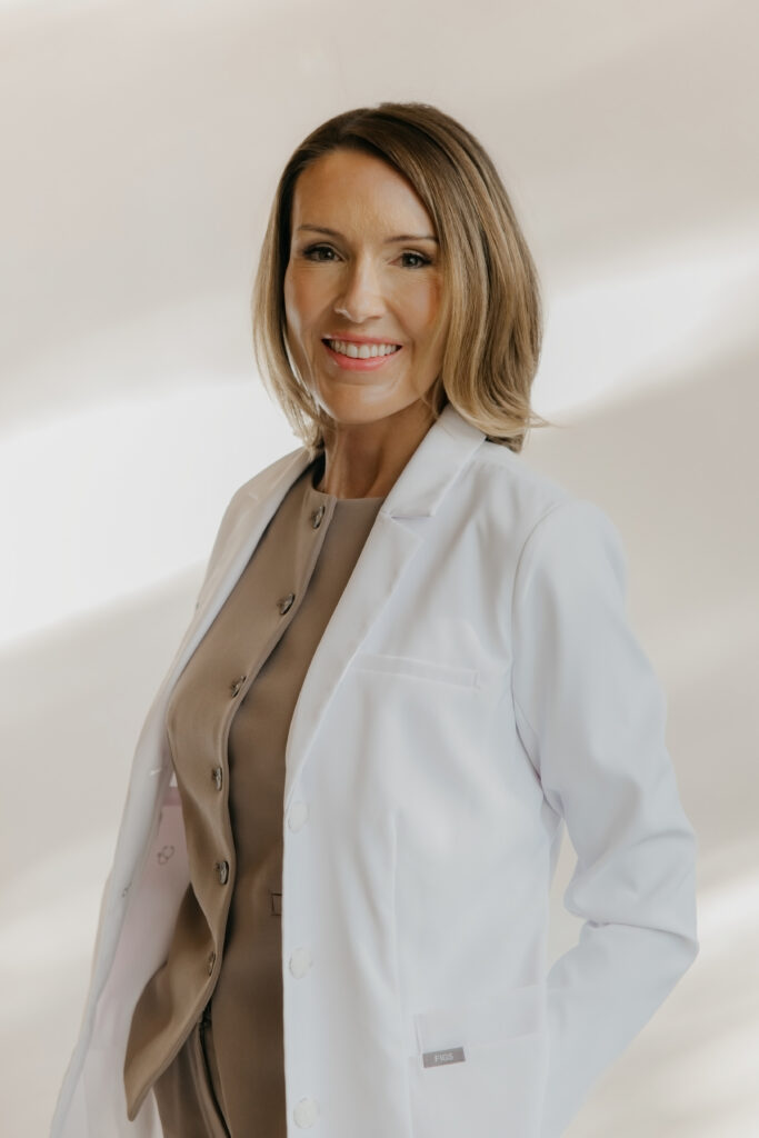 Stephanie Astor in a white lab coat over a tan dress, smiling with hand on hip, nurse injector brand photography