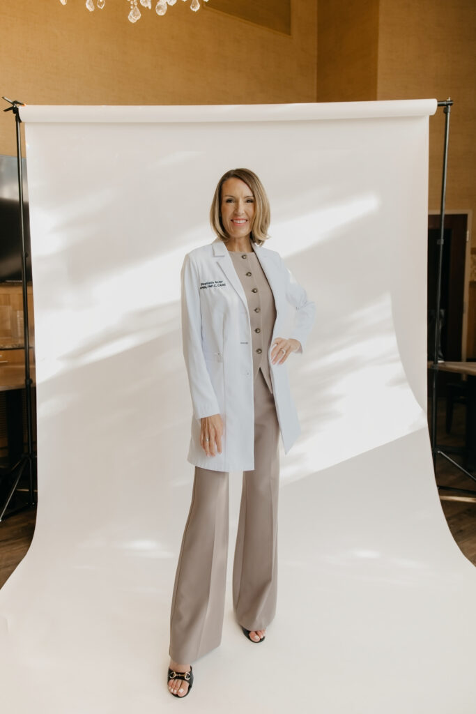 Stephanie Astor standing in a white lab coat over a taupe outfit against a white studio backdrop, full-body brand photo