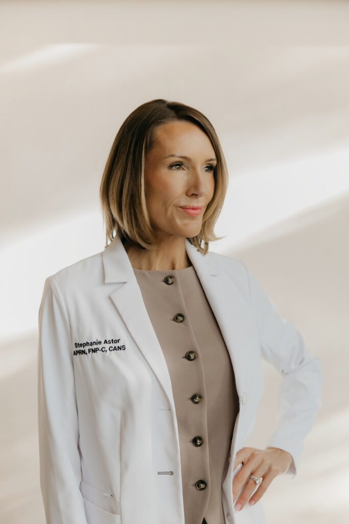 Stephanie Astor standing in a white lab coat over a beige top, hand on hip, side profile