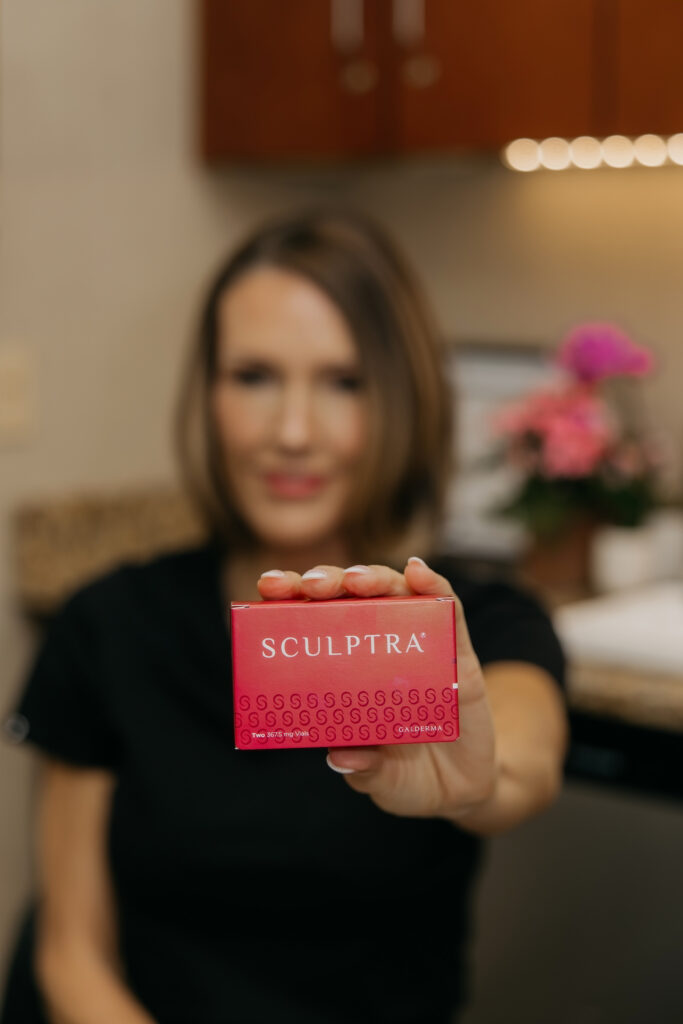 Stephanie Astor smiling while holding a Sculptra box toward the camera, pink flowers in background