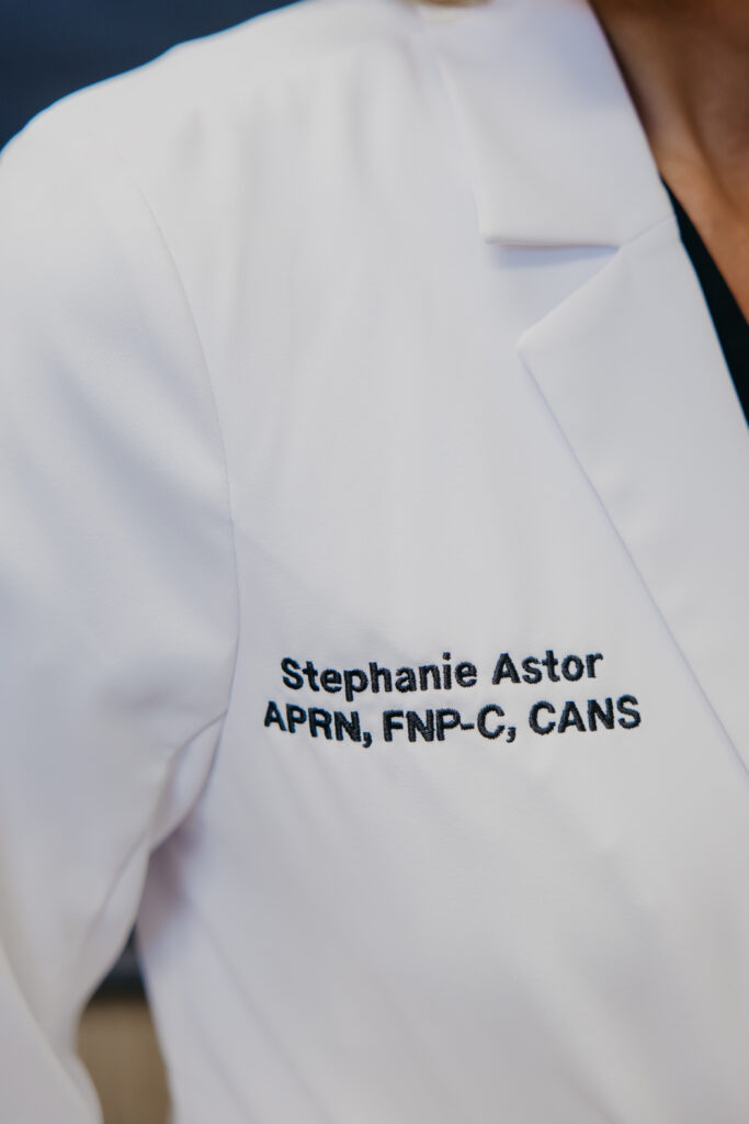 Close-up of a white coat lapel embroidered "Stephanie Astor, APRN, FNP-C, CANS"