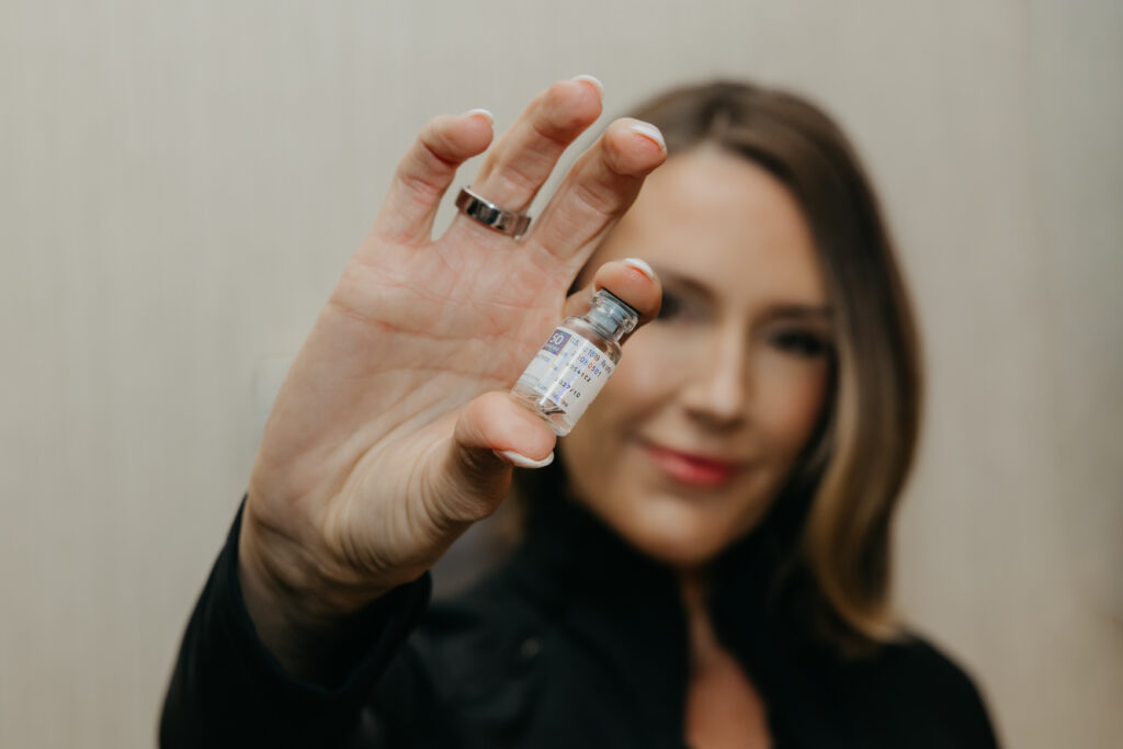 Close-up of Stephanie Astor's face softly blurred behind a filler vial held toward the camera