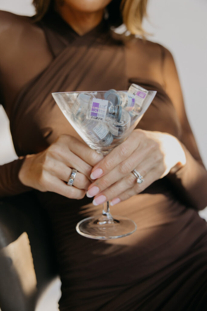 Hands in a brown sleeve arranging injectable vials inside a martini glass, styled product shot