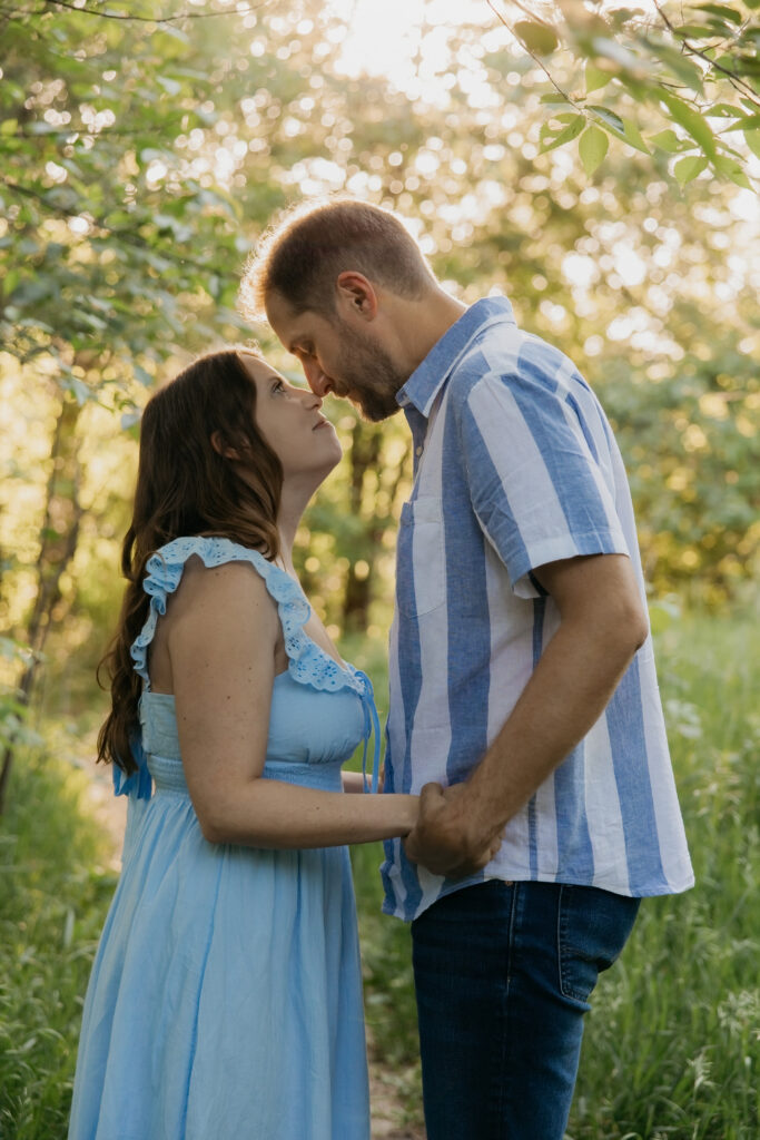 Couple nose to nose holding hands on a wooded path, woman in light blue dress