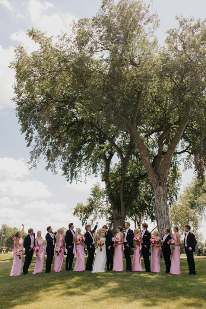 Full wedding party standing together under a large shade tree