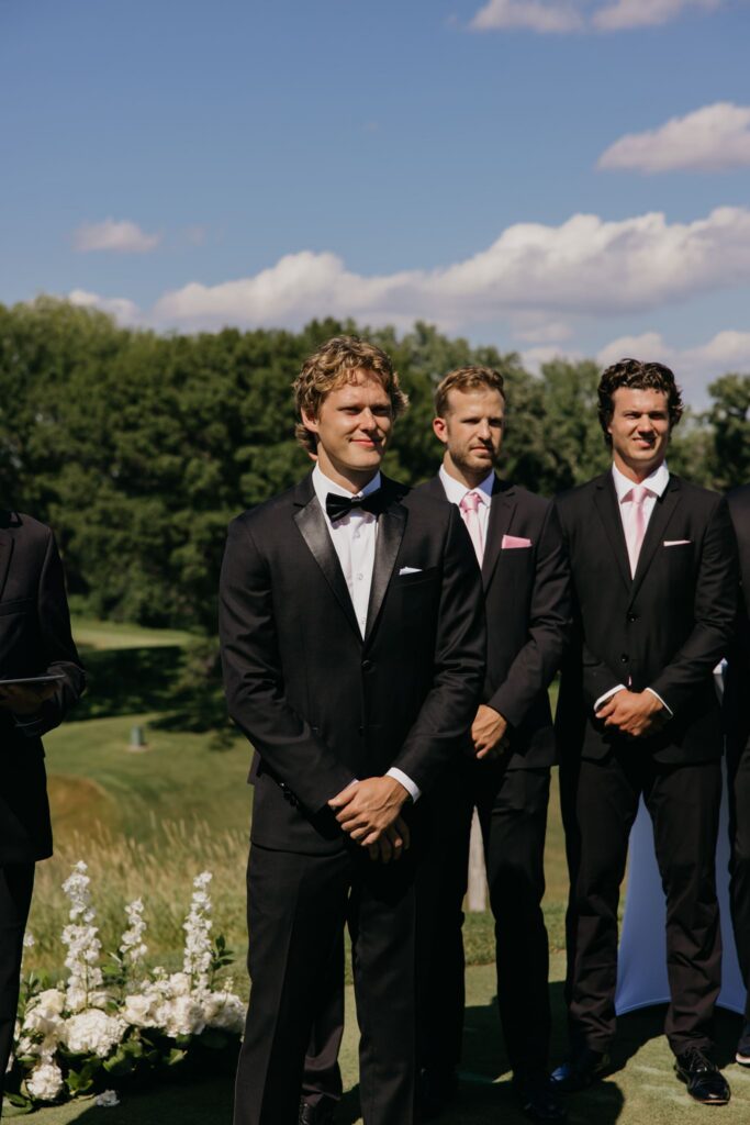 Groomsmen in matching black tuxedos and pink ties standing during the ceremony