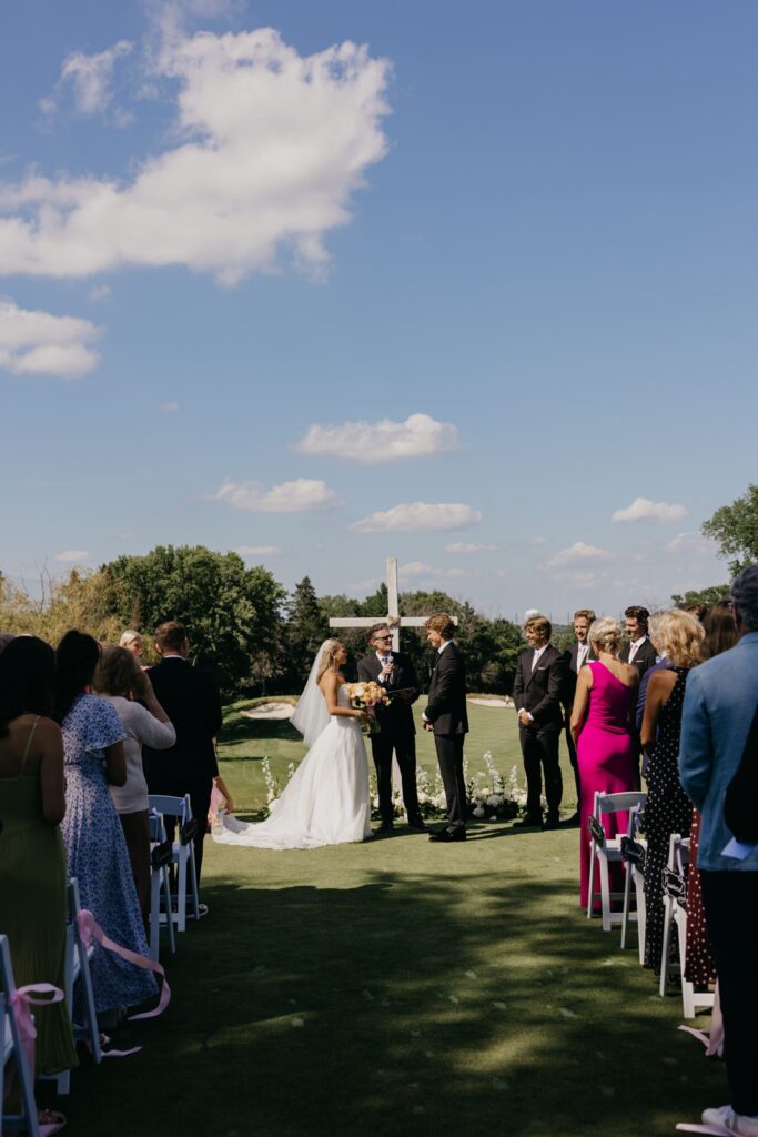 Guests seated for outdoor wedding ceremony with white cross and golf course backdrop