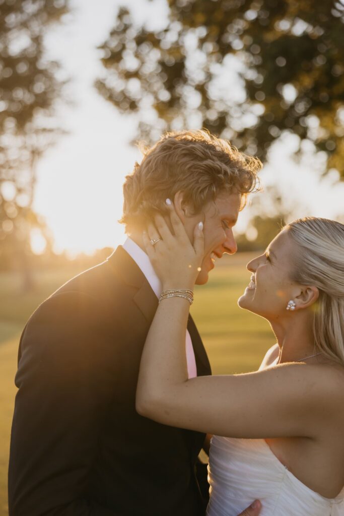 Groom laughing as bride touches his face during golden hour portraits on the golf course