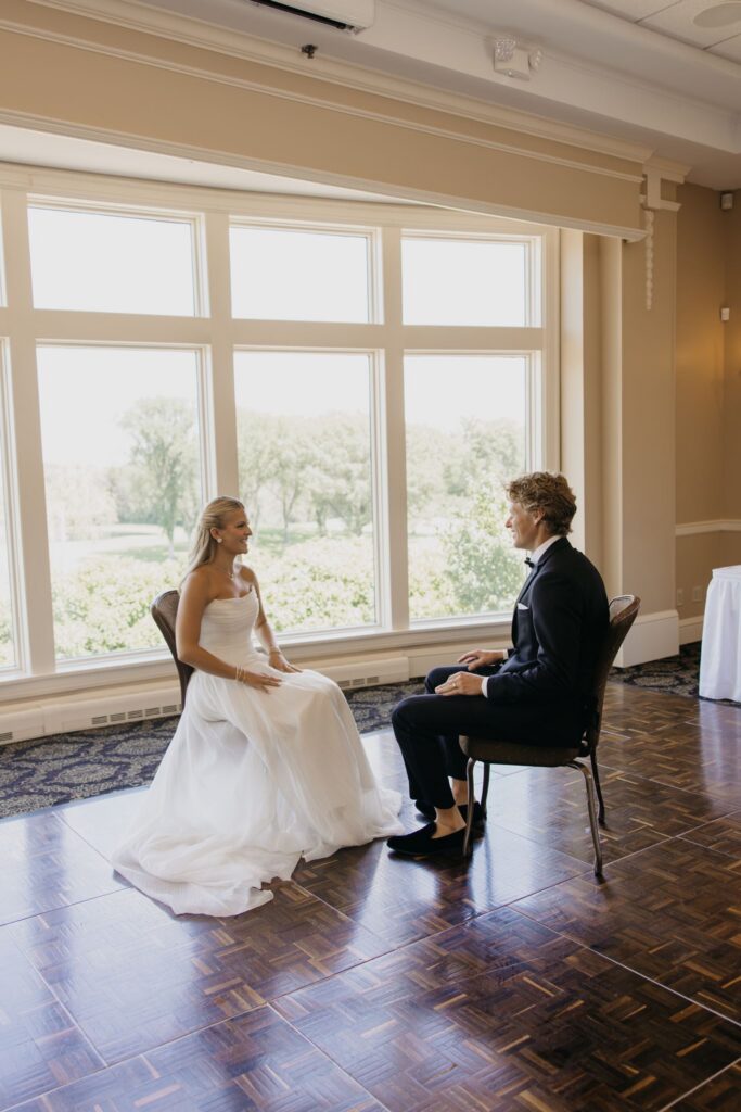 Bride and groom seated by large windows during their first look reaction