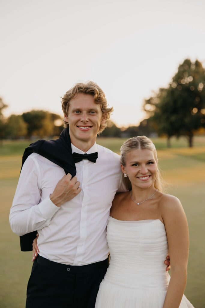 Groom smiling with jacket over shoulder standing beside bride on the golf course at sunset
