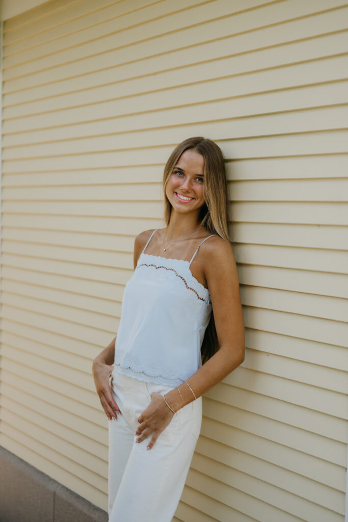 Senior girl in a white cami and pants leans against a house exterior with hand on hip