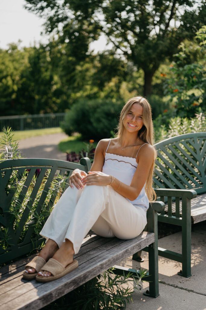 Senior girl in a white cami and pants sits on a green garden bench