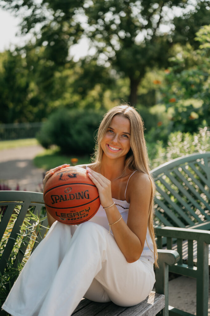 Senior girl in white outfit sits on a green garden bench holding a basketball