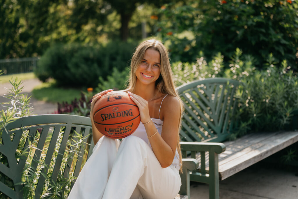 Close-up of senior girl in white outfit smiling on a garden bench while holding a basketball