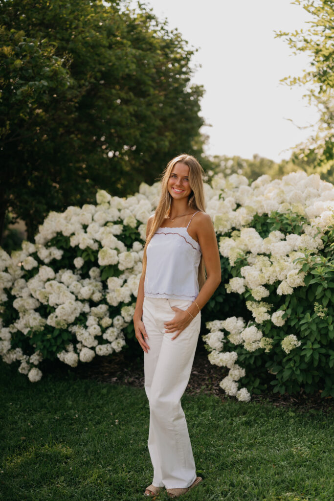 Senior girl in a white cami and pants stands beside a blooming white hydrangea bush