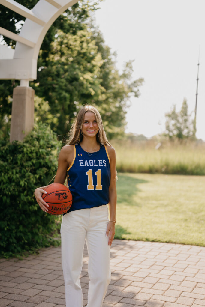Senior girl in blue Eagles jersey stands under a garden pergola holding a basketball, wide shot