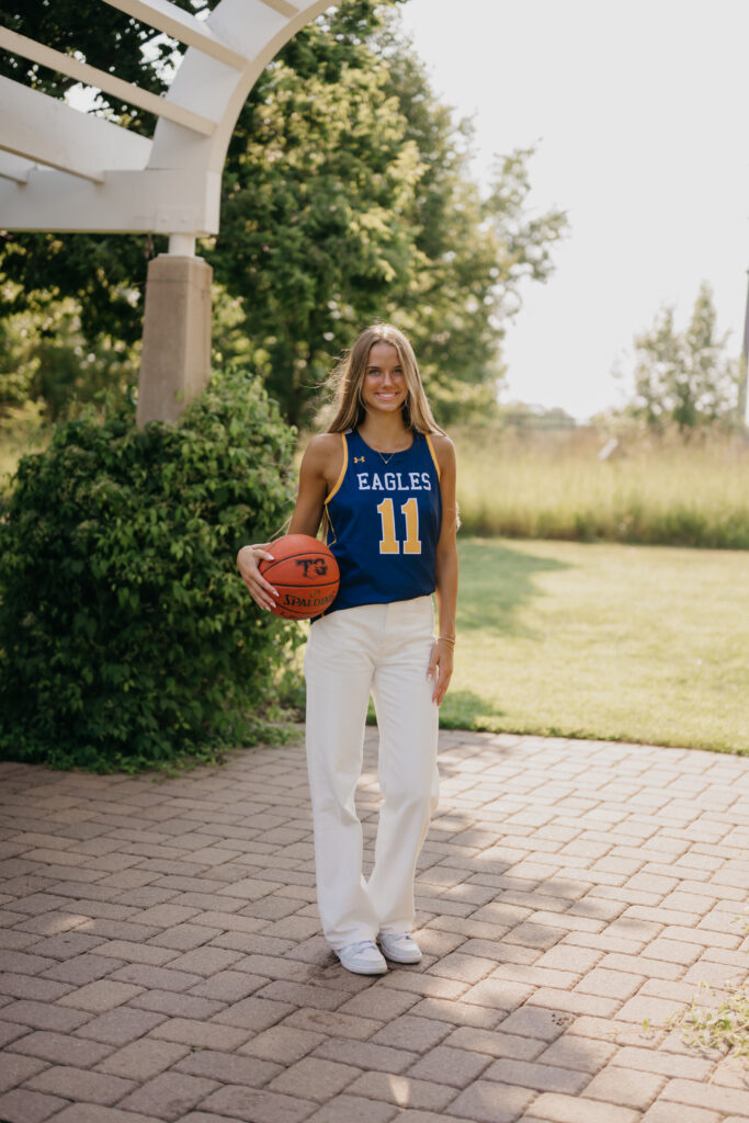 Senior girl in blue Eagles jersey stands under a pergola holding a basketball, wide shot