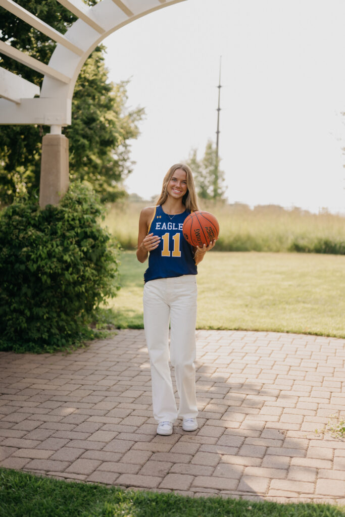 Senior girl in blue Eagles jersey stands on a brick garden path holding a basketball