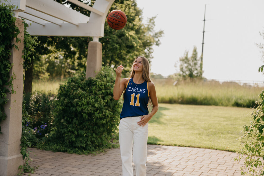 Senior girl in blue Eagles jersey tosses a basketball into the air, looking up under a pergola