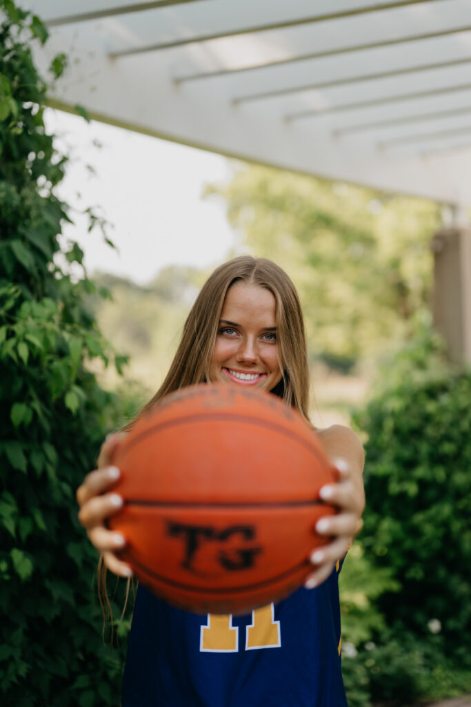 Close-up of senior girl in blue Eagles jersey holding a basketball toward the camera under a garden pergola