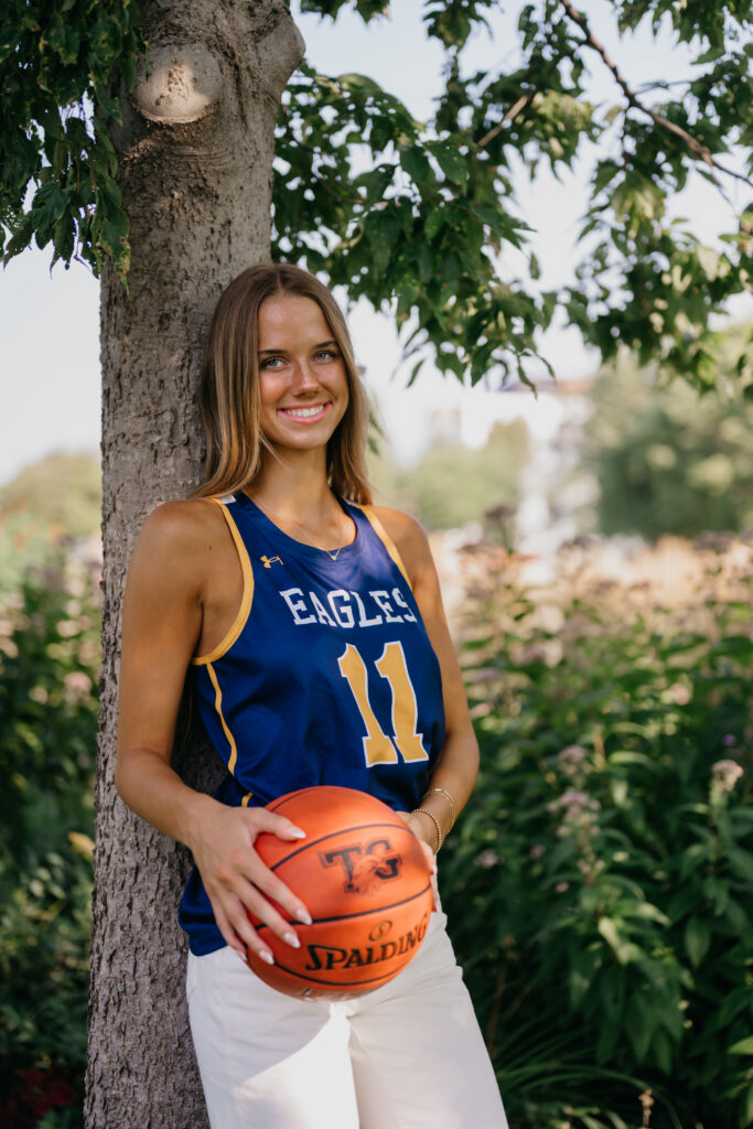 Senior girl in blue Eagles jersey leans against a tree trunk holding a basketball close to her chest