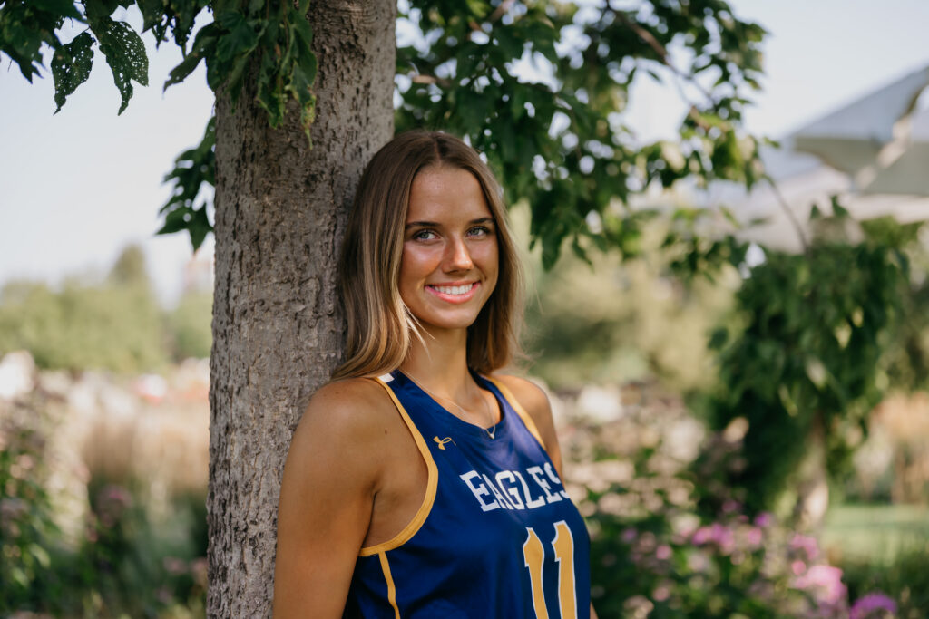 Senior girl in blue Eagles basketball jersey leans against a tree trunk in a garden for her athlete senior photos