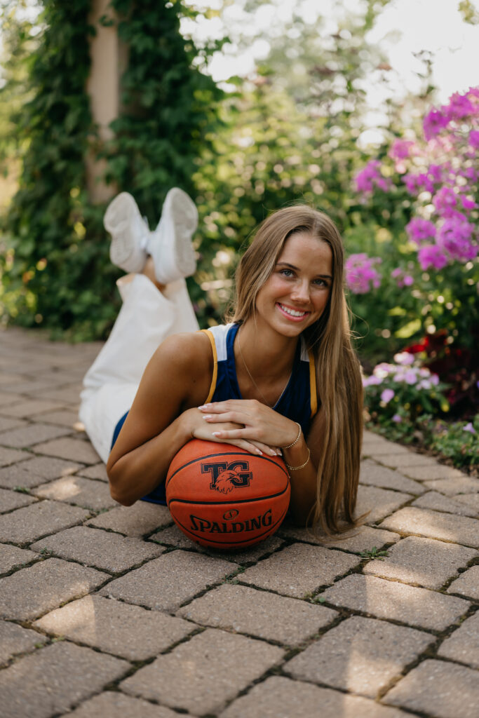 Senior girl in blue Eagles jersey lying on the patio, resting on a basketball, side angle