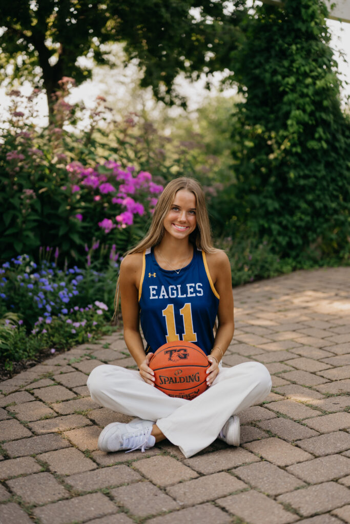 Senior girl in blue Eagles basketball jersey sits cross-legged on a brick patio, holding a basketball, with purple flowers behind her