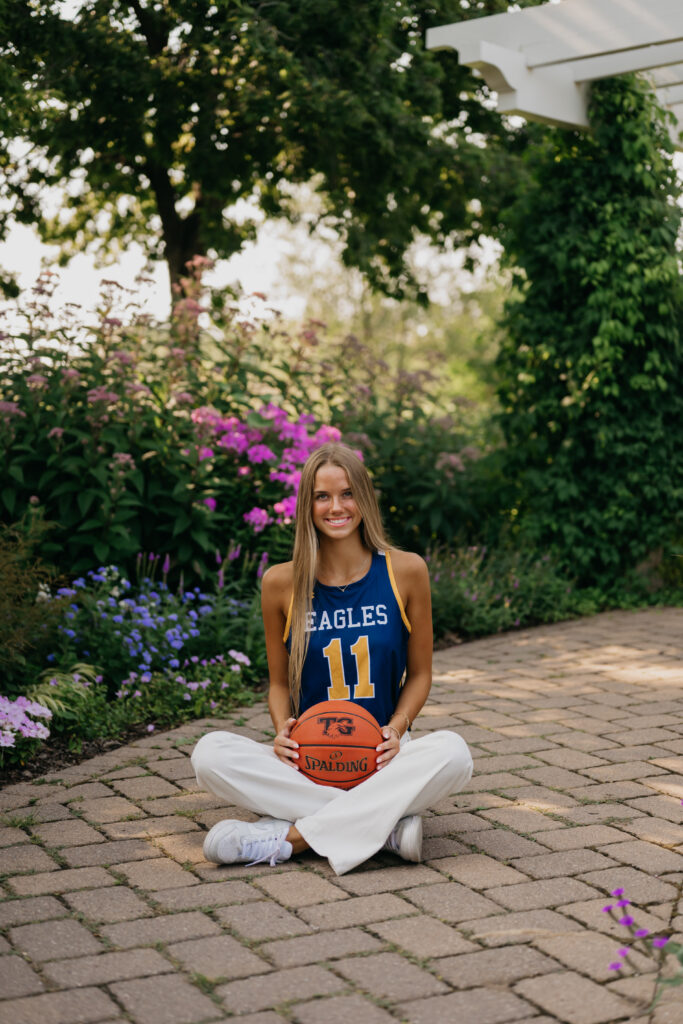 Senior girl in blue Eagles jersey sits cross-legged on a brick patio with a basketball on her lap, flowers behind her
