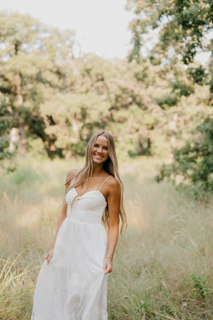 Senior girl in a flowing cream dress stands in a sunlit grass field