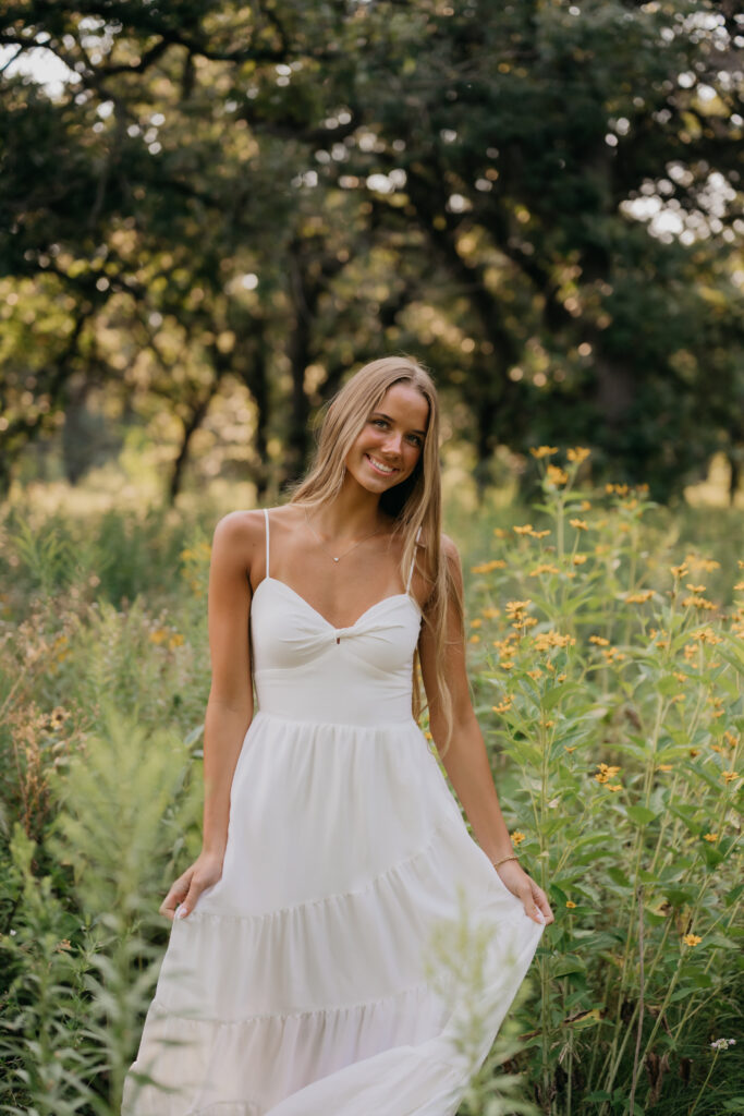 Senior girl in a white tiered maxi dress holds her skirt while standing in a wildflower field