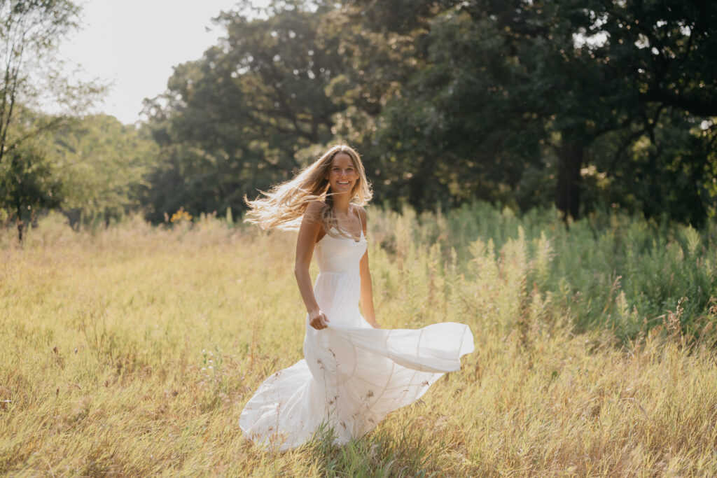 Senior girl in a white dress walks through a wildflower field, hair blowing, skirt in motion