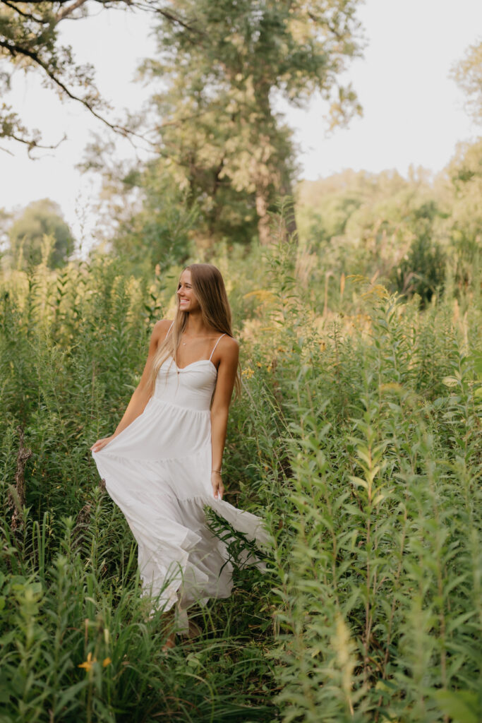 Senior girl in a flowing white maxi dress walks through a tall grass field
