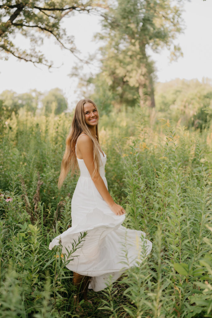 Senior girl in a white dress twirls through tall grass, skirt in motion