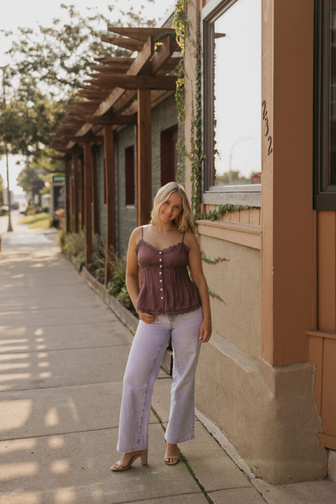 Senior girl standing on a sidewalk near a storefront in a mauve top and jeans