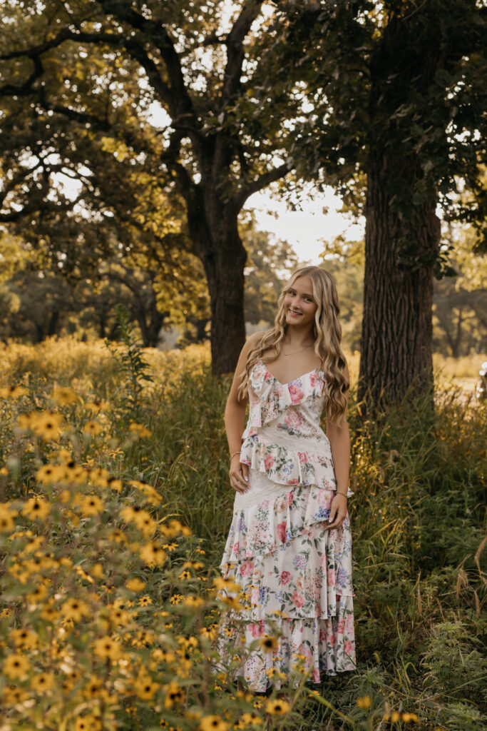 Senior girl standing under oak trees in a wildflower field in a floral maxi dress