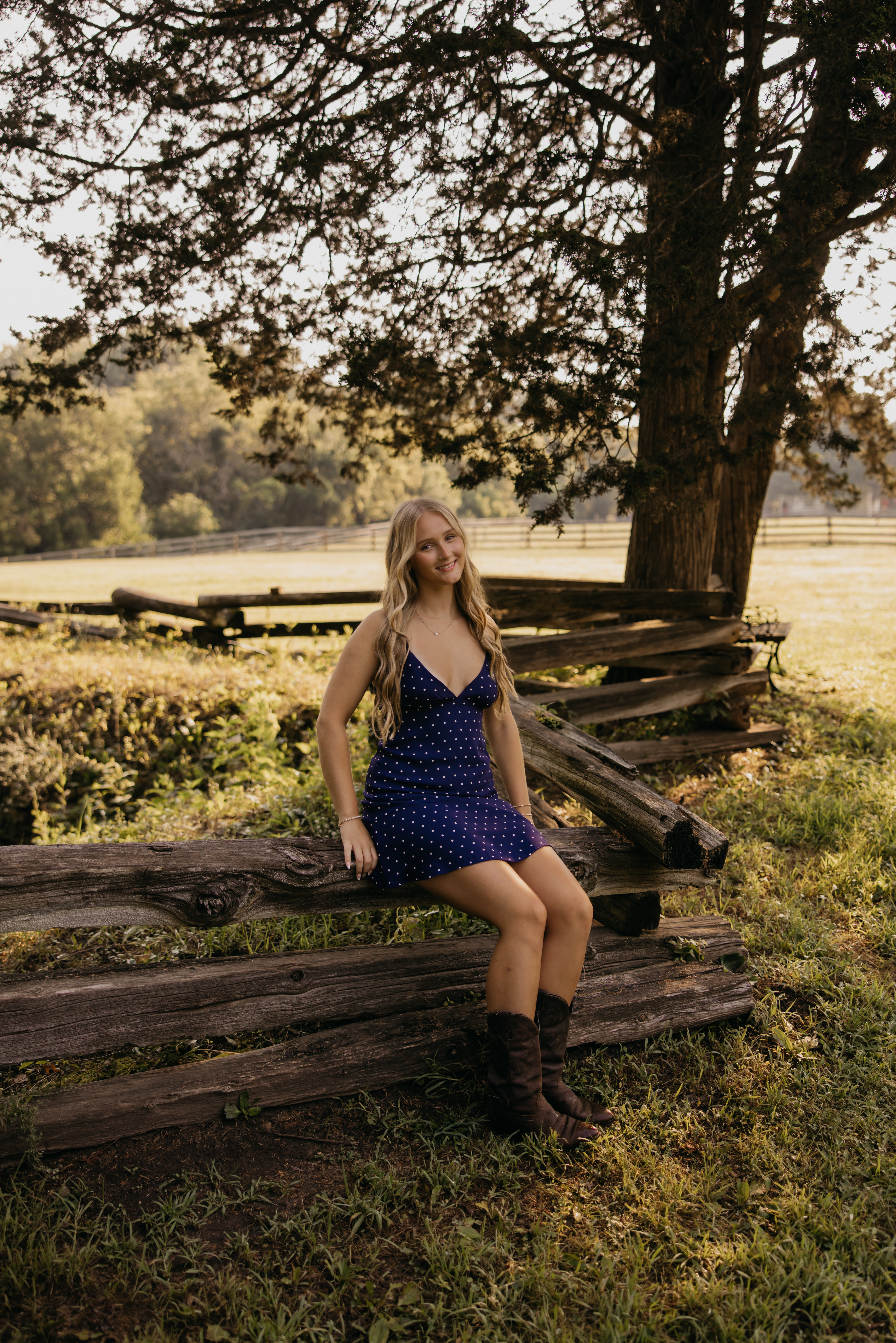 Senior girl in a navy polka-dot dress sitting on a wooden split-rail fence for a romantic senior pictures Minneapolis session