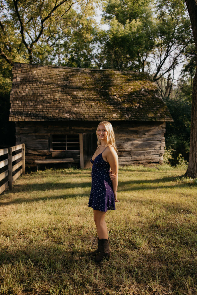 Senior girl standing in tall grass near a weathered wood cabin